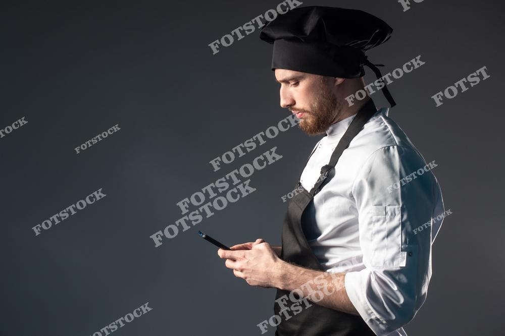 На фотографии мужчина в поварской униформе с черным колпаком и фартуком, который сосредоточенно использует смартфон. Темный фон подчеркивает современный подход к кулинарии, где цифровые устройства становятся частью рабочего процесса.